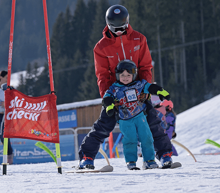 Skilehrer führt ein kleines Kind mit Helm und Startnummer zwischen den Beinen über die Piste