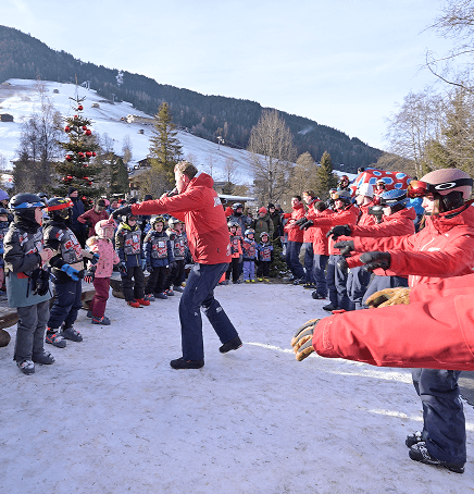 Skilehrer in roten skiCHECK-Jacken tanzen beim Aufwärmen mit einer großen Kindergruppe im Schnee