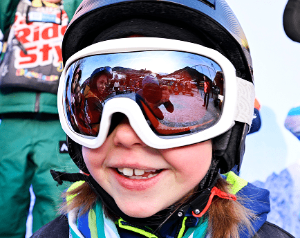 Lachendes Kind mit weißer Skibrille, in der sich das Skischulgelände spiegelt
