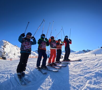 Kindergruppe in Skikurs-Leibchen streckt auf der Piste die Skistöcke in die Luft