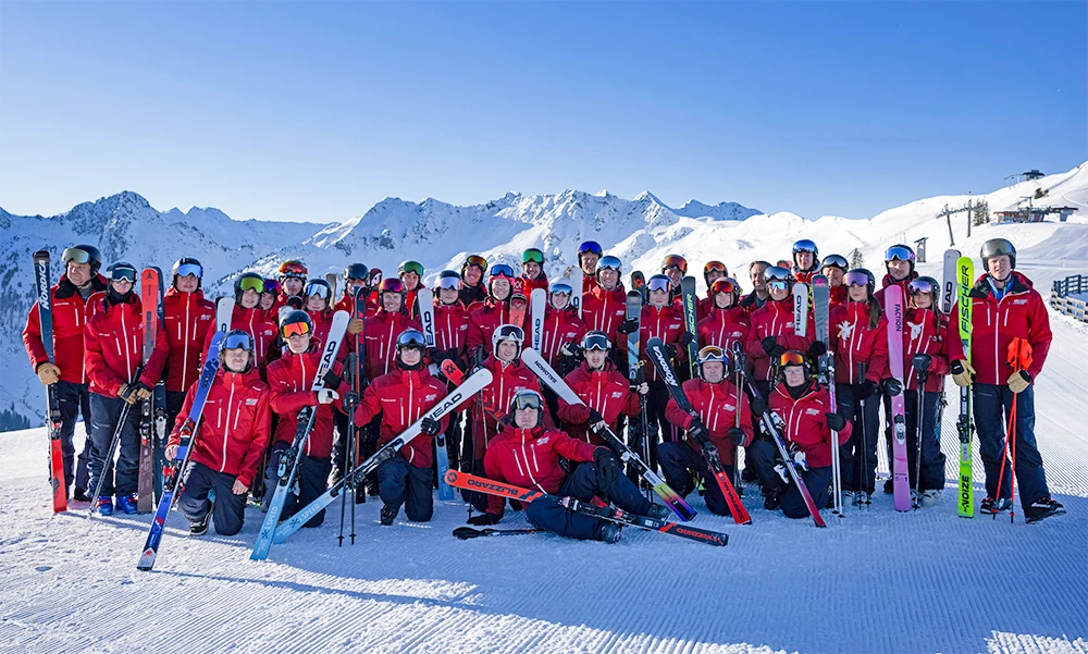 Gruppenbild des skiCHECK-Skilehrerteams in roten Jacken mit Ski vor verschneitem Bergpanorama