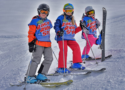 Drei Kinder in Race-Ride-Style-Leibchen stehen mit Ski auf der Piste