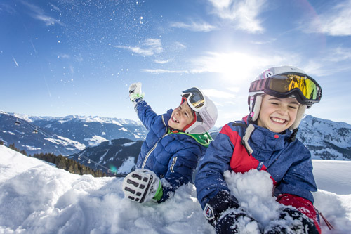 Zwei lachende Kinder spielen mit Schnee vor einem sonnigen Bergpanorama
