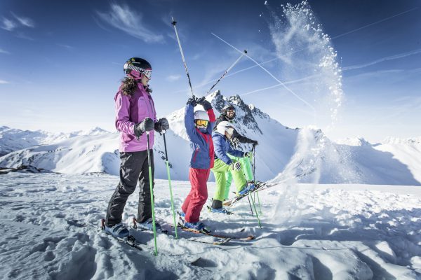 Familie mit Ski auf dem Berggipfel, ein Kind wirbelt Schnee in die Luft