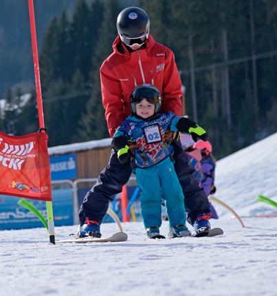 Skilehrer führt ein kleines Kind mit Helm und Startnummer zwischen den Beinen über die Piste
