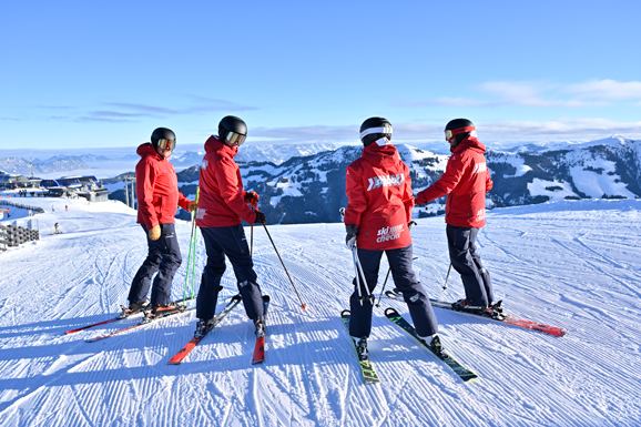 Vier skiCHECK-Skilehrer in roten Jacken stehen auf der Piste und blicken auf das Bergpanorama