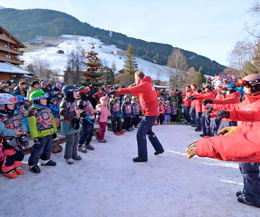 Skilehrer in roten skiCHECK-Jacken tanzen beim Aufwärmen mit einer großen Kindergruppe im Schnee
