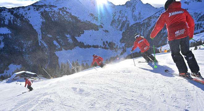 skiCHECK-Skilehrer in roten Jacken fahren eine sonnige Piste vor verschneiten Bergen hinab