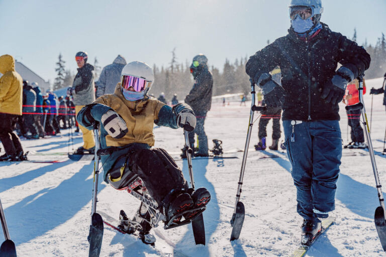 Person im Monoski mit Krückenskiern neben einem Begleiter mit Stützskiern auf sonniger Piste